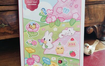 Un livre d'autocollants intitulé « Cozy Stickers », dont la couverture est ornée d'adorables illustrations représentant un lapin, un poussin, un gâteau, une fraise et des fleurs. Le livre est posé sur une surface en bois, à côté d'une petite figurine d'oiseau.